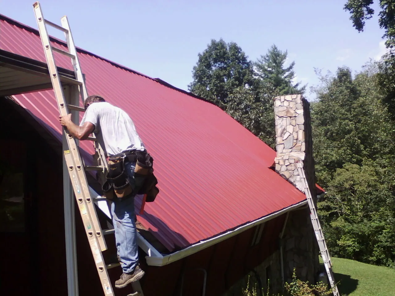 Roofer on a ladder beside a red metal roof and stone chimney — Soffit & Fascia Repair by Roofing Brothers in Kahlotus